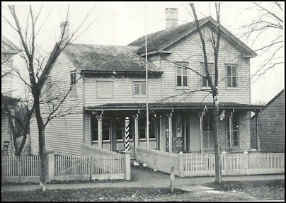 A historical black and white photograph of a two-story wooden house with a front porch and decorative elements, surrounded by trees and a picket fence.