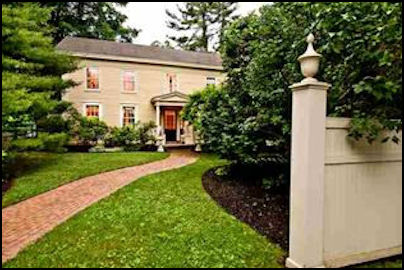A charming two-story house with a welcoming front porch, surrounded by lush greenery and well-maintained landscaping. A paved pathway leads to the entrance, framed by a white fence and gate.