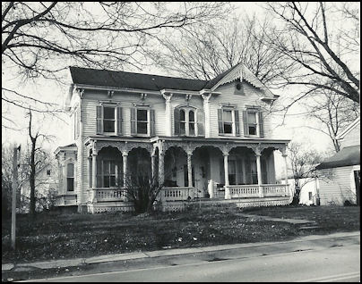 Black and white photograph of an old Victorian house with intricate woodwork, large front porch, and multiple windows, surrounded by trees.