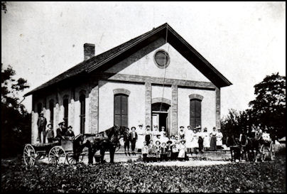 Historic black and white photo of a large group of people gathered outside a schoolhouse, featuring a horse-drawn wagon and surrounded by greenery.
