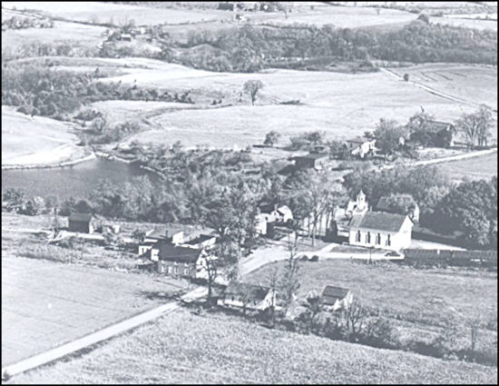 Aerial view of a small rural community featuring a church and several houses surrounded by fields and a river.