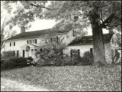 Black and white photograph of a traditional house surrounded by trees and fallen leaves, showcasing a front porch and multiple windows.