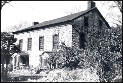 Black and white photograph of an old stone house surrounded by greenery.